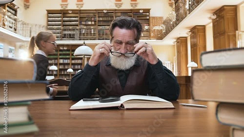Tired senior bearded man taking off his glasses and massaging the temples feeling a headache beacause his noisy grandchildren interfering to read book in the library photo
