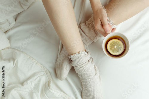 Cozy flatlay of woman's legs in warm white stockings in bed holding cup of lemon tea, selective focus