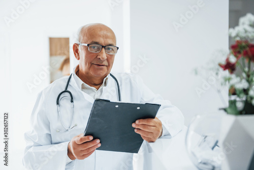 Portrait of senior doctor with notepad and in white uniform that stands in the clinic