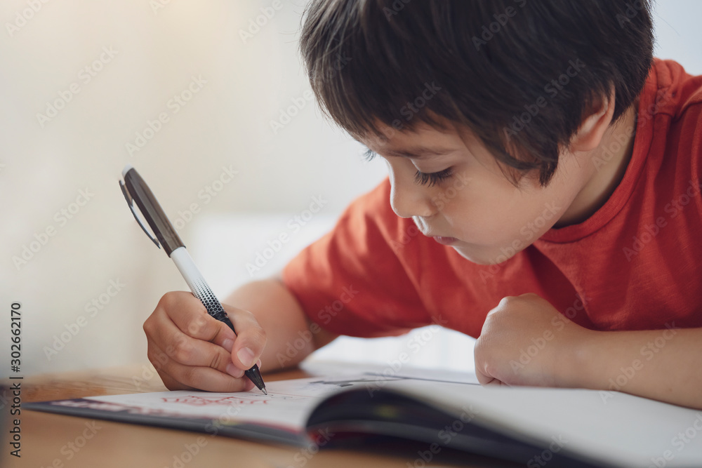 Selective focus of school kid boy siting on table doing homework, Happy Child holding black pen,Little boy is writing on white paper, Elementary school, Homeschooling concept Selective focus of school kid boy siting on table doing homework, Happy Child holding black pen,Little boy is writing on white paper, Elementary school, Homeschooling concept