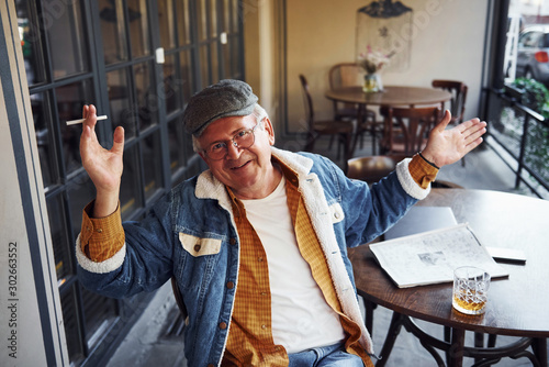 Stylish senior in fashionable clothes and in glasses sits in the cafe with cigarette and talks