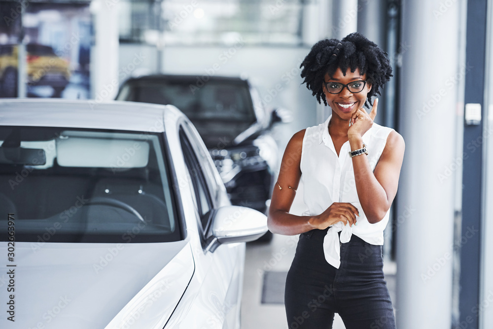 Young african american woman in glasses stands in the car salon near vehicle Young african american woman in glasses stands in the car salon near vehicle
