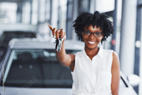 Fotografia Young african american woman in glasses stands in the car salon near vehicle wit