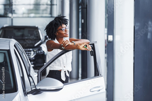 Foto Young african american woman in glasses stands in the car salon near vehicle wit