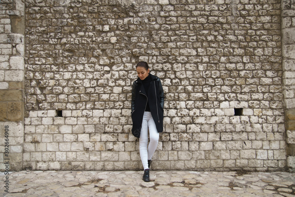 Young woman dressed in white jeans and black coat stands leaning on an ancient wall made of stone blocks and looks down