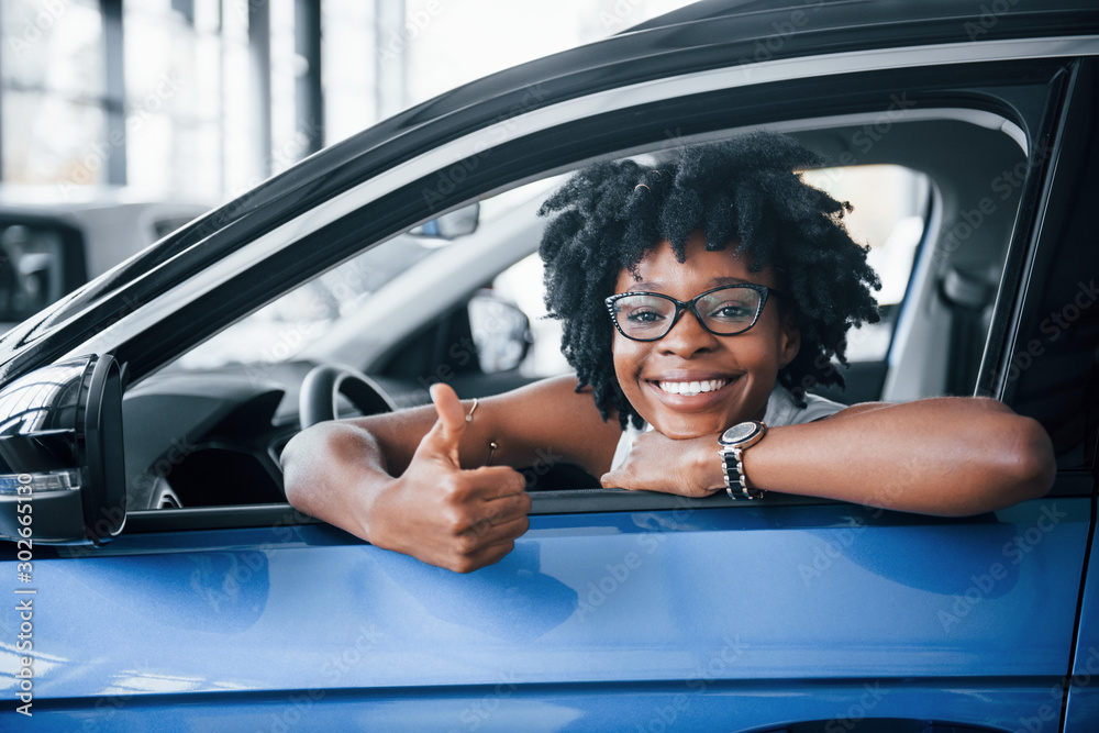 Shows thumb up. Young african american woman sits inside of new modern car - obrazy, fototapety, plakaty 