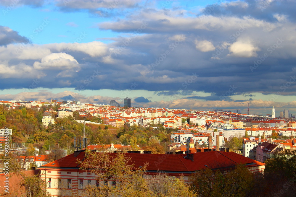 Panorama Of Prague. Houses with orange roofs, under sunlight, among yellowed trees against a blue sky with heavy clouds.