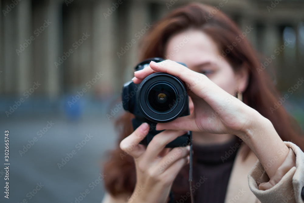 Selective focus on lens. Beautiful stylish fashionable girl holds camera in her hands and takes pictures. Woman photographer with long dark hair in city, urban shoot, vertical. unrecognizable person