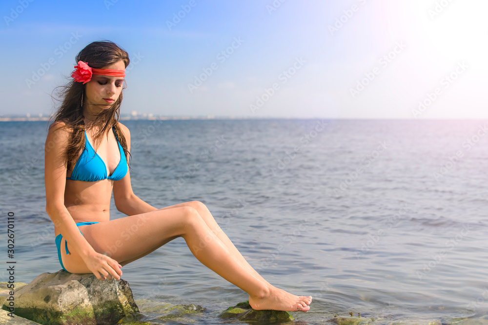 young brunette girl in a bright blue bathing suit sits on a large stone  against the sea. Copy space фотография Stock | Adobe Stock