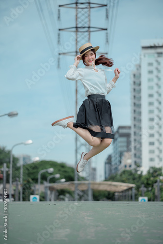 Cheerful pretty asian girl in white shirt black skirt and boater hat jumping for joy with hand on hat on a beautiful blue sky day her face is full of happiness © KsDear