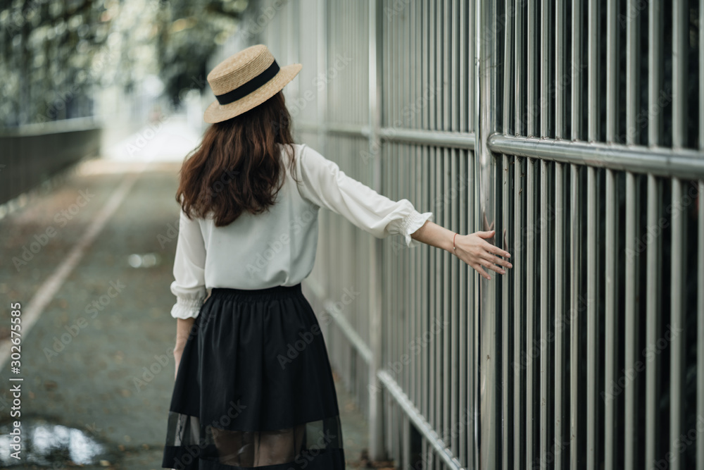 asian woman with long wavy hair in white shirt and boater hat walking on green pathway turned her back to the camera and touching the fence on the right side enjoying moment while walking home alone