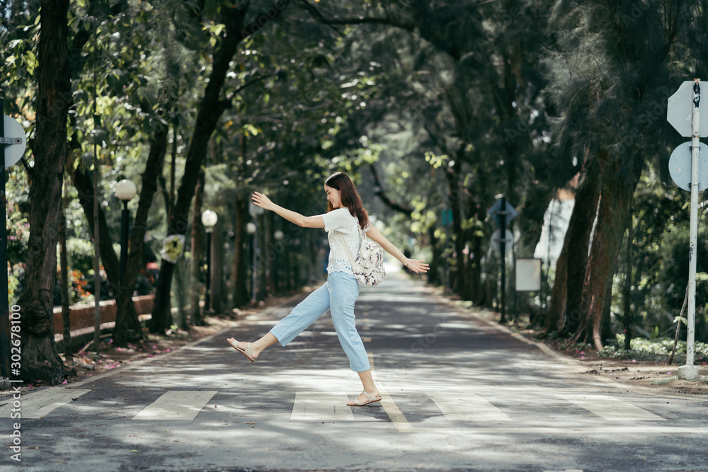 young woman is happy, lady is smiling, woman in white t shirt has happiness, happy asian girl with backpack stepping and swinging arms while crossing street surrounded by tree tunnel 
