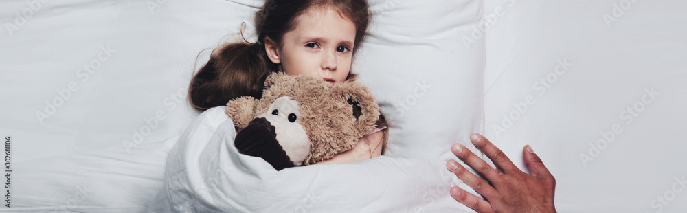 partial view of male hand near scared child lying in bed with teddy bear, panoramic shot