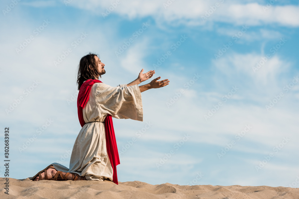 jesus praying on knees on sand in desert against sky - obrazy, fototapety, plakaty 