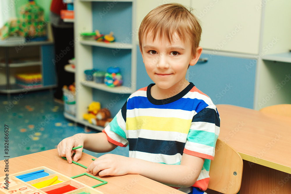 Fototapeta premium Happy boy sits at the desk at classroom. Kid using colorful sticks and learning counting. Education, development and school concept