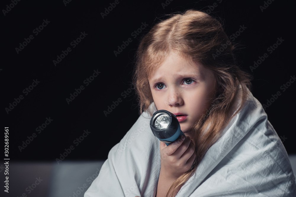 Fototapeta premium scared kid sitting under blanket and holding flashlight isolated on black