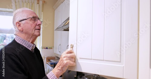 Forgetful Senior Man With Dementia Looking In Cupboard At Home