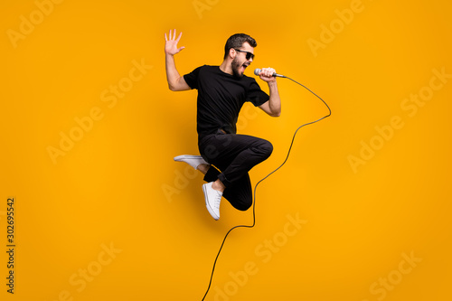 Full body profile photo of crazy hipster guy jumping high holding microphone music lover singing favorite song wear sun specs black t-shirt pants isolated yellow color background photo