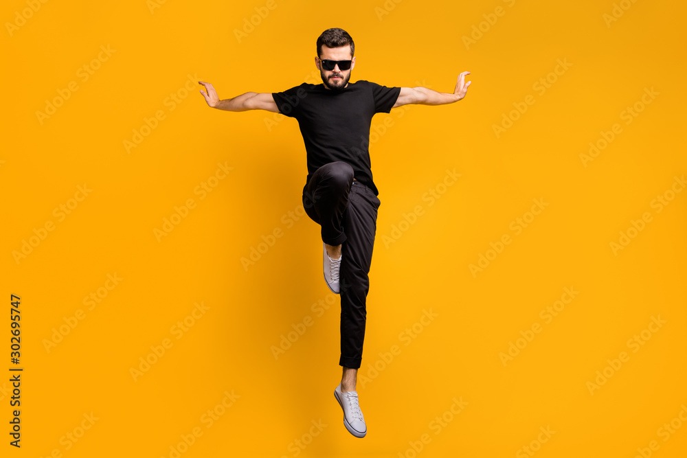 Full body photo of crazy guy jumping high practicing self defense kicking confident facial expression wear sun specs black t-shirt pants isolated yellow color background