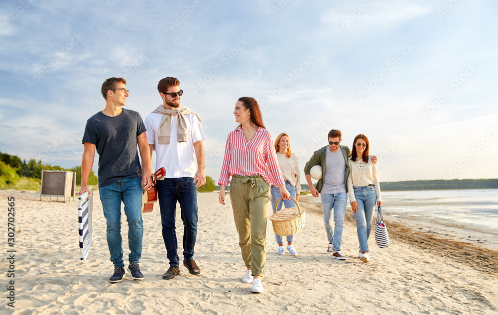 friendship, leisure and people concept - group of happy friends with ball, guitar, bag and picnic basket with blanket walking along beach in summer