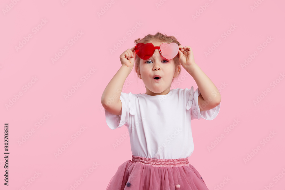 Portrait of surprised cute little toddler girl in the heart shape sunglasses. Child with open mouth having fun isolated over pink background. Looking at camera. Wow funny face - obrazy, fototapety, plakaty 