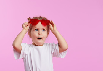 Portrait of surprised cute little toddler girl in the heart shape sunglasses. Child with open mouth having fun isolated over pink background. Looking at camera. Wow funny face