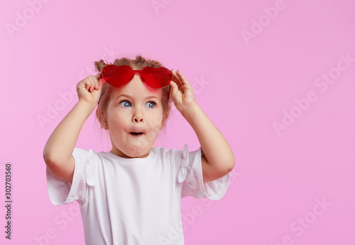 Obraz na płótnie Portrait of surprised cute little toddler girl in the heart shape sunglasses. Child with open mouth having fun isolated over pink background. Looking at camera. Wow funny face w salonie