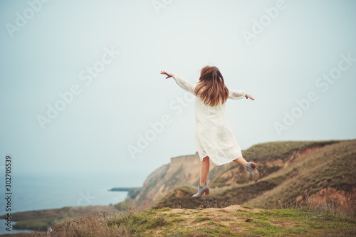 Dreams come true! Success woman in white dress at sunset or sunrise jumping and flying with arms raised above her head in celebration of having reached mountain top summit goal.