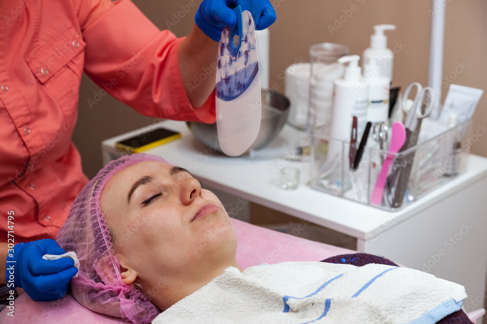 Naklejka premium A young girl is lying on a couch during cosmetic procedures with a mask on the face above which beautician woman сools the skin with a fan from burning. Cleansing and make up.