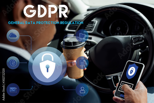 back view of african american businessman using smartphone with gdpr illustration and drinking coffee in car