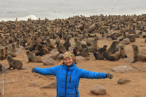 Young Caucasian red-haired happy girl traveler backpacker in a blue down jacket with spread out arms against the background of a huge colony of fur seals in Cape Kros in Namibia, Africa. photo