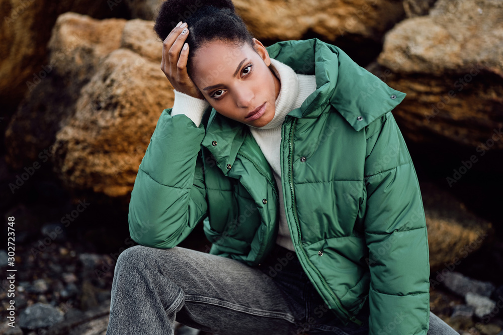 Obraz premium Portrait of casual African American girl in stylish down jacket intently looking in camera while posing on stones by the sea