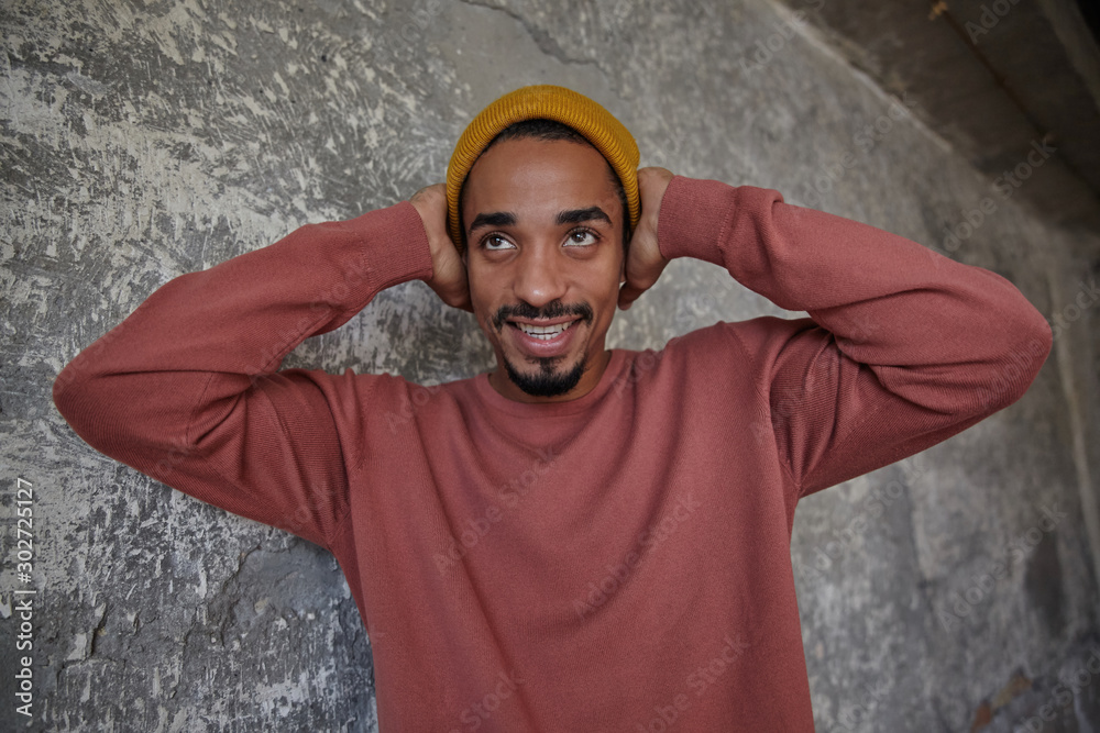 Photo of positive young dark skinned man with brown eyes covering his ears with raised palms and smilling cheerfully while looking aside, having fun with his friends over concrete background Photo of positive young dark skinned man with brown eyes covering his ears with raised palms and smilling cheerfully while looking aside, having fun with his friends over concrete background