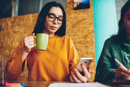 Pensive student with brunette hair and eyeglasses reading notification on smartphone while enjoying tasty tea during break.Female office manager with mug of beverage checking email on celluler photo