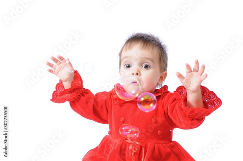 Funny surprised little girl in red Christmas dresses plays with soap bubbles on white isolated background. Happy childhood