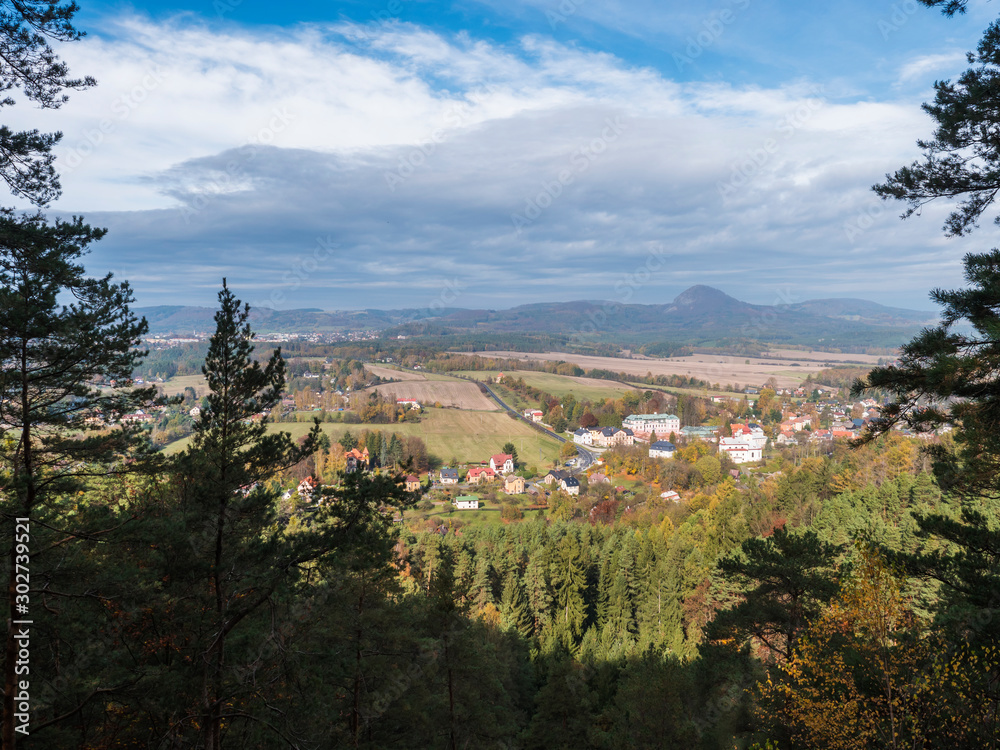 View from Hrabencina vyhlidka lookout on village Sloup v cechach in luzicke hory, Lusatian Mountains with autumn colored deciduous and coniferous tree forest and green hills, blue sky, white clouds