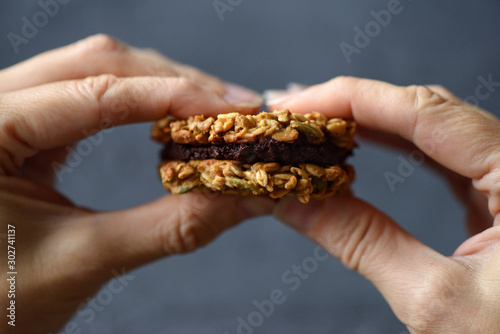 Dieting or low calorie food concept. Close up of woman hands holding oatmeal breakfast cookie sandwich with dark chocolate stuffing on black background