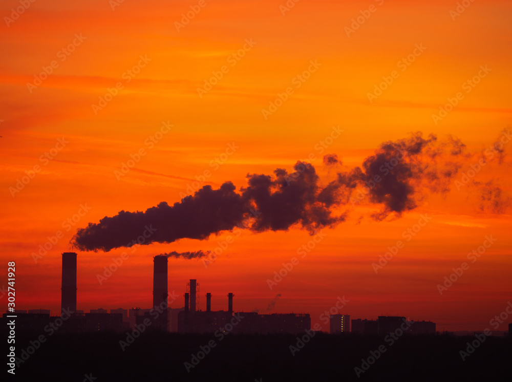 Tall smoking chimneys from industrial plant and far buildings behind the dark forest. Cityscape silhouette with colorful twilight sky on sunset. Moscow city, Russia.