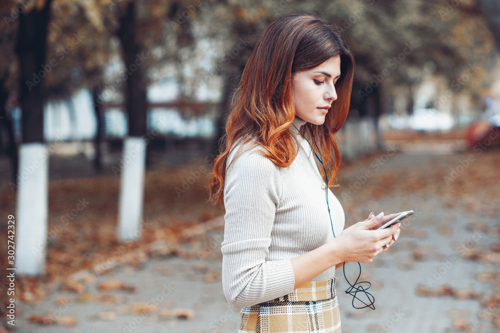 Image of beautiful stylish woman holding mobile phone. Young european girl standing at the street and using cellphone. Woman listening to music with the phone and having fun. Mobile internet concept. Image of beautiful stylish woman holding mobile phone. Young european girl standing at the street and using cellphone. Woman listening to music with the phone and having fun. Mobile internet concept.