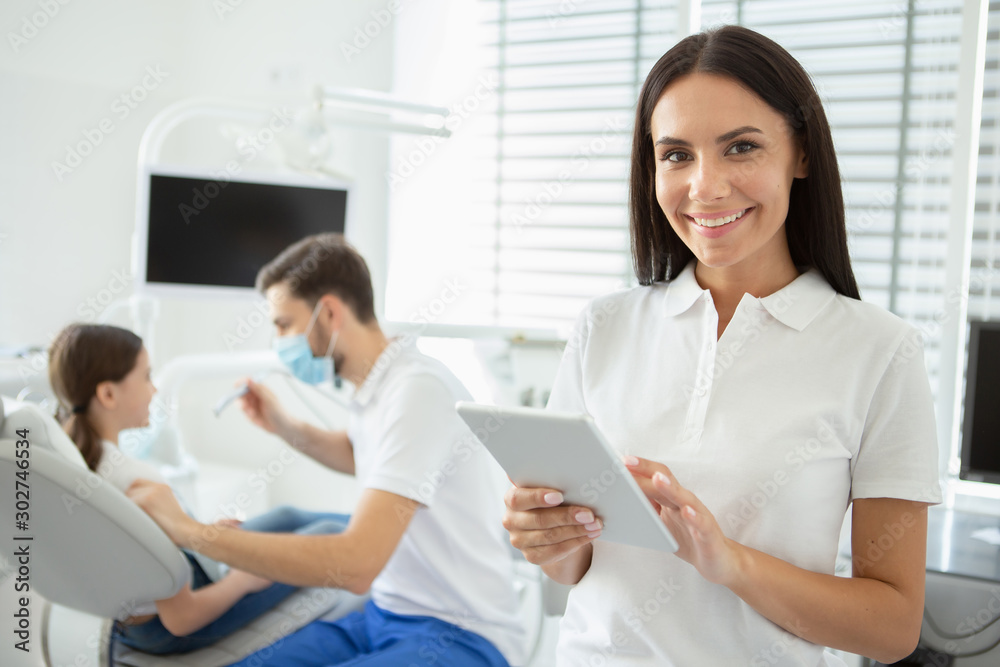 smiling dentist working on digital tablet and looking at the camera with male doctor working with client on the background smiling dentist working on digital tablet and looking at the camera with male doctor working with client on the background