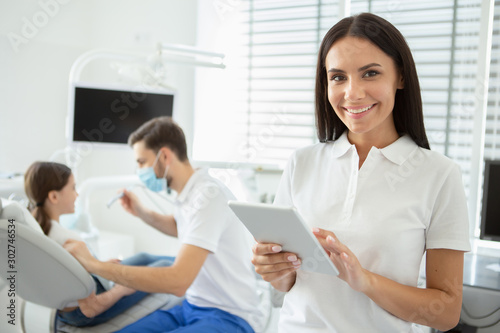 smiling dentist working on digital tablet and looking at the camera with male doctor working with client on the background
