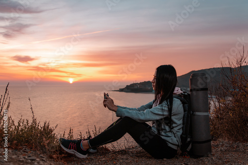 The concept of Hiking and sports recreation. A woman with a backpack on her back sits on a rock and looks at the sunset  holding a mobile phone. In profile. Copy space