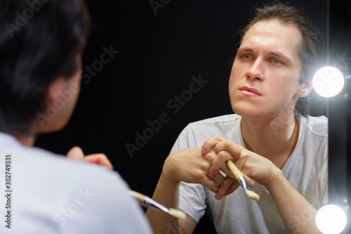 the actor sits in front of a mirror in a white t-shirt in a dark room and looks at his reflection, holding a brush for applying makeup to his face