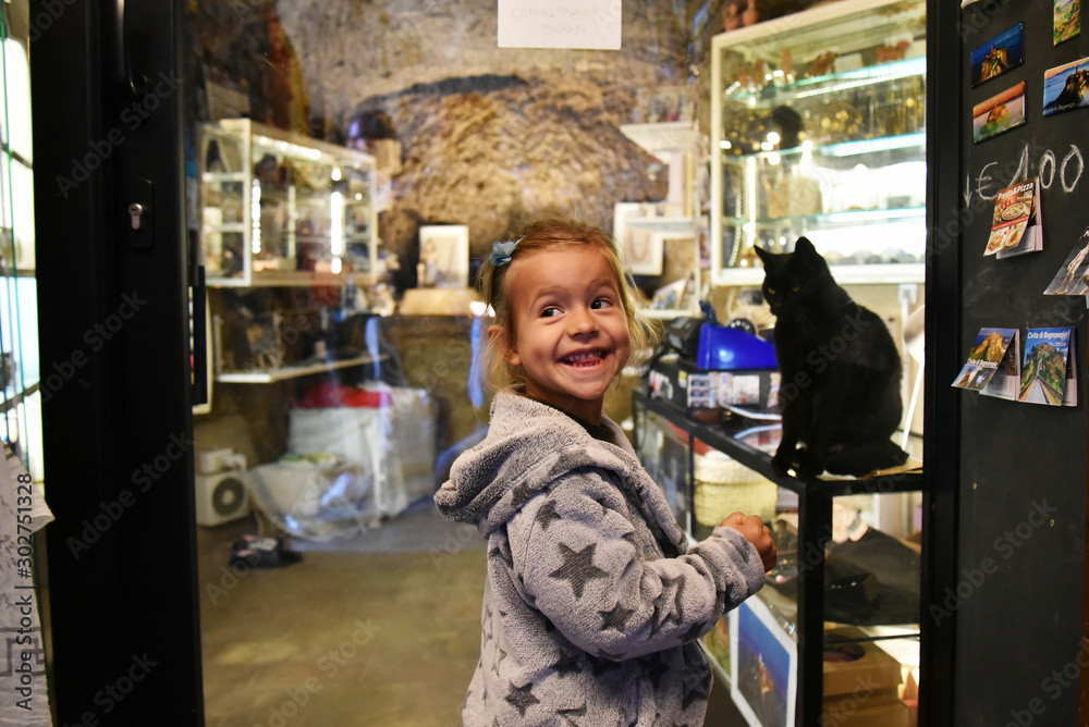 Civita di Bagnoregio, Lazio, Italy - October 10, 2019:  Funny little girl in front of a souvenir shop. In the shop there is a black cat.