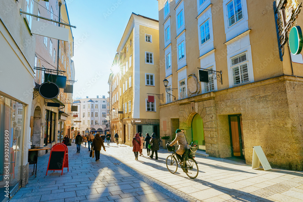 Naklejka premium Kobieta na rowerze na ulicy starego miasta Salzburga w Austrii. Turyści i ludzie w mieście Mozart, Europa, zima. Panorama i punkt orientacyjny. Pejzaż miejski z rozbłyskiem słońca. Sklepy i sklep. Architektura budynku