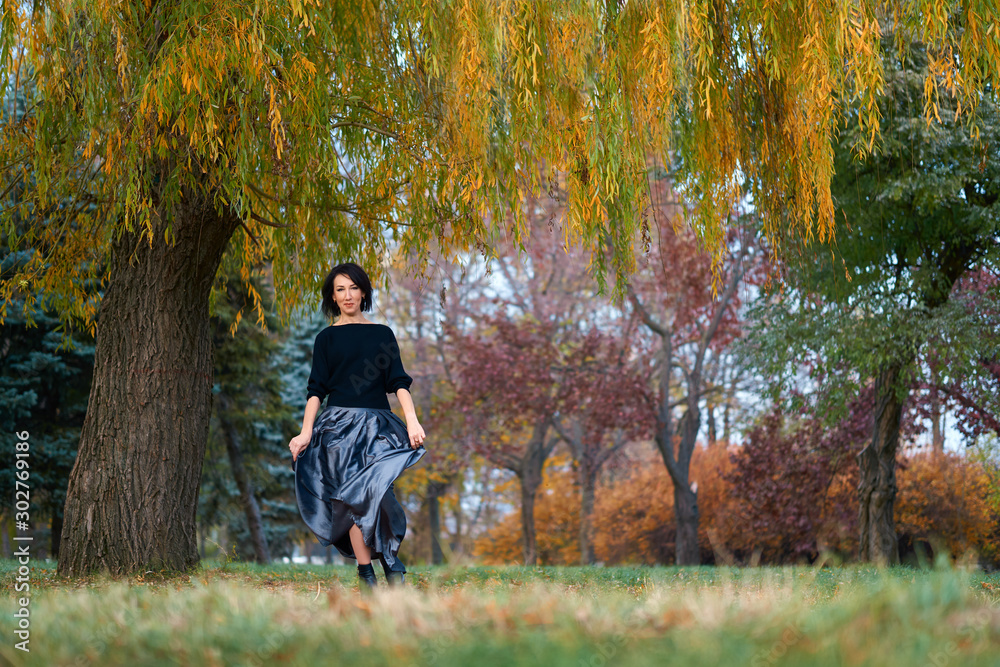 Beautiful elegant woman standing and posing near big yellow tree in a city park in autumn