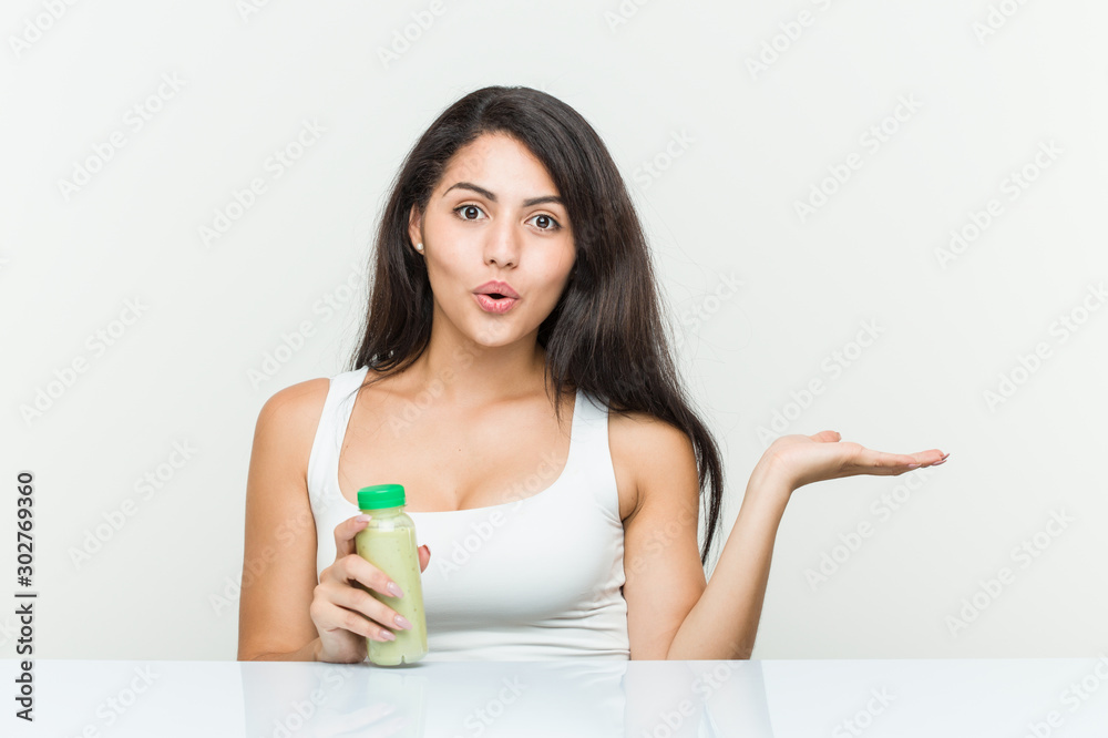 Young hispanic woman holding a vegetable drink impressed holding copy space on palm. Young hispanic woman holding a vegetable drink impressed holding copy space on palm.