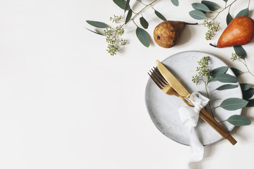 Autumn thanksgiving table place setting. Golden cutlery, porcelain plate, berry eucalyptus leaves and branches, silk ribbon and pear fruit on white table background. Fall wedding. Flat lay, top view.