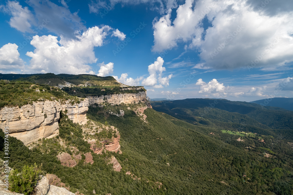 High rock with blue sky and white clouds. Green forest at the bottom of the valley, Tavertet, Catalonia.