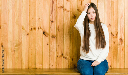 Young chinese woman sitting on a wooden place tired and very sleepy keeping hand on head.